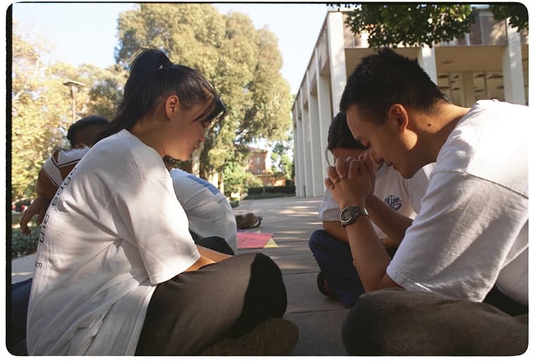 A group seated outdoors with heads bowed and hands clasped