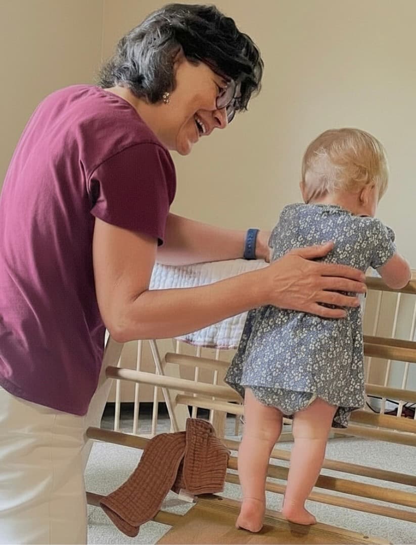 A woman steadies a standing toddler with both hands.