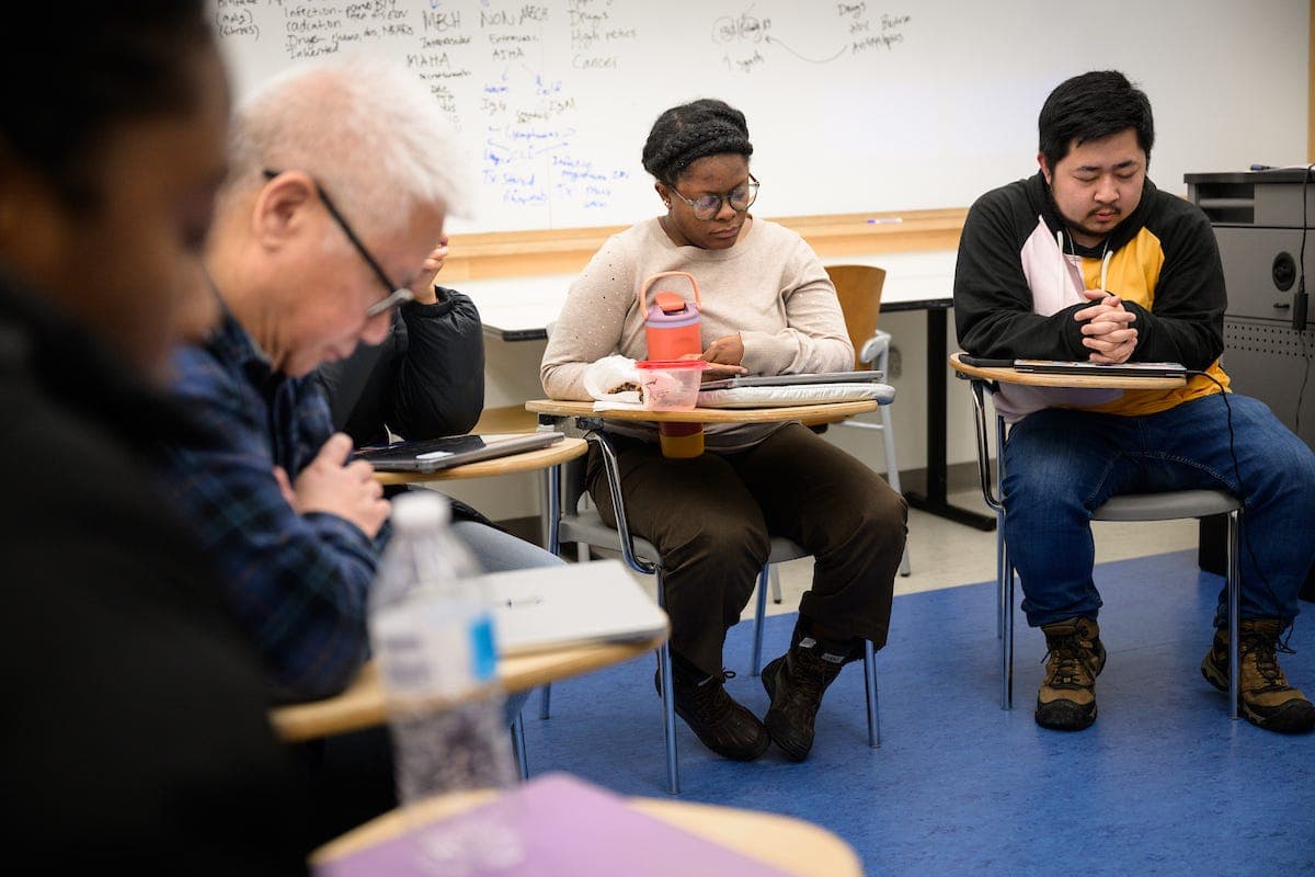 Several people sit in a classroom, reading and writing at desks.