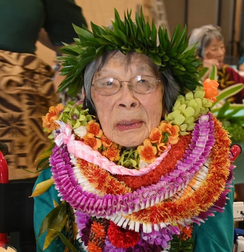 A woman wears glasses, a leafy head wreath, and several colorful flower leis.