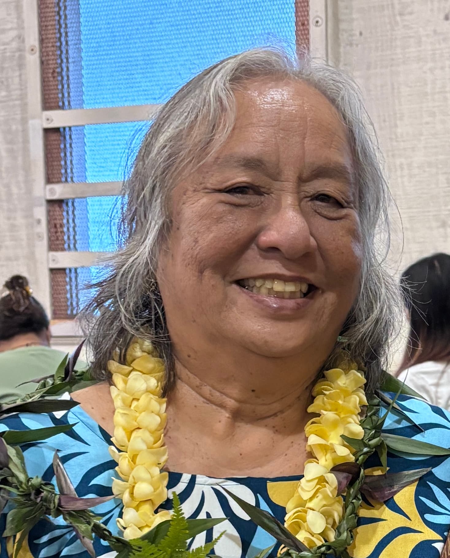 A smiling woman with gray hair wears a floral shirt and a yellow flower lei.