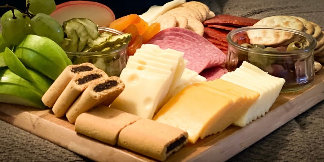 A wooden serving board arranged with cheeses, sliced meats, crackers, and jars of condiments.