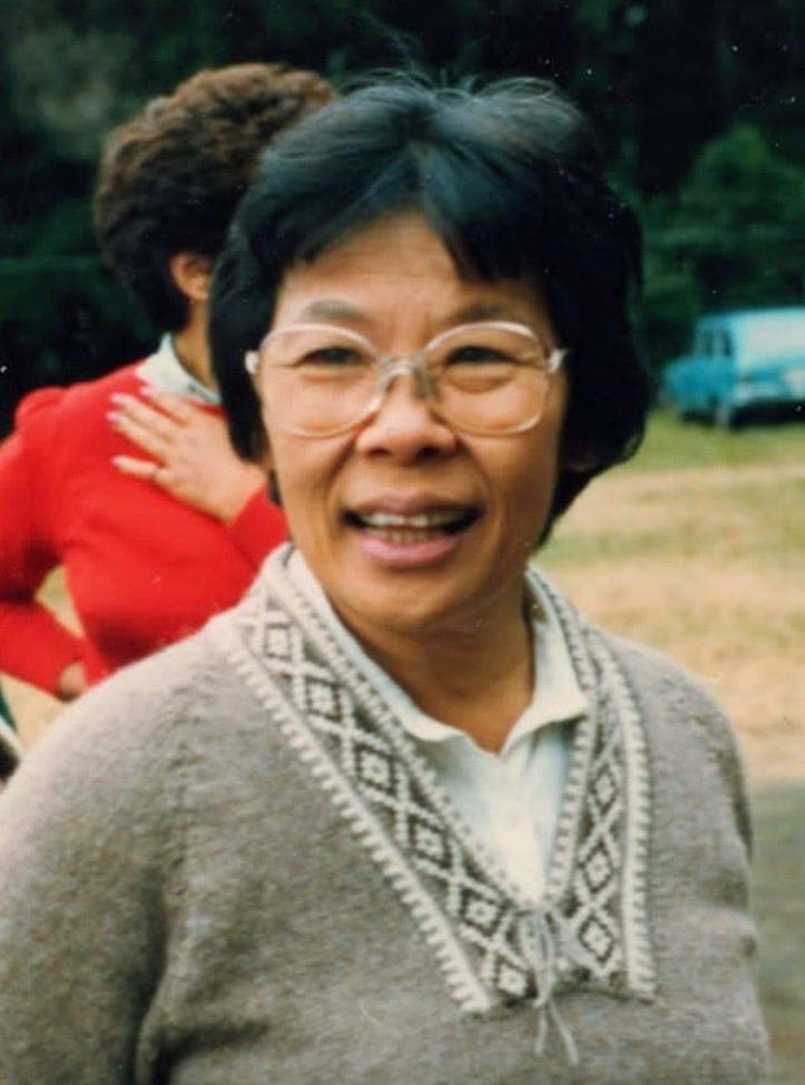 A smiling woman wearing glasses and a gray cardigan outdoors.