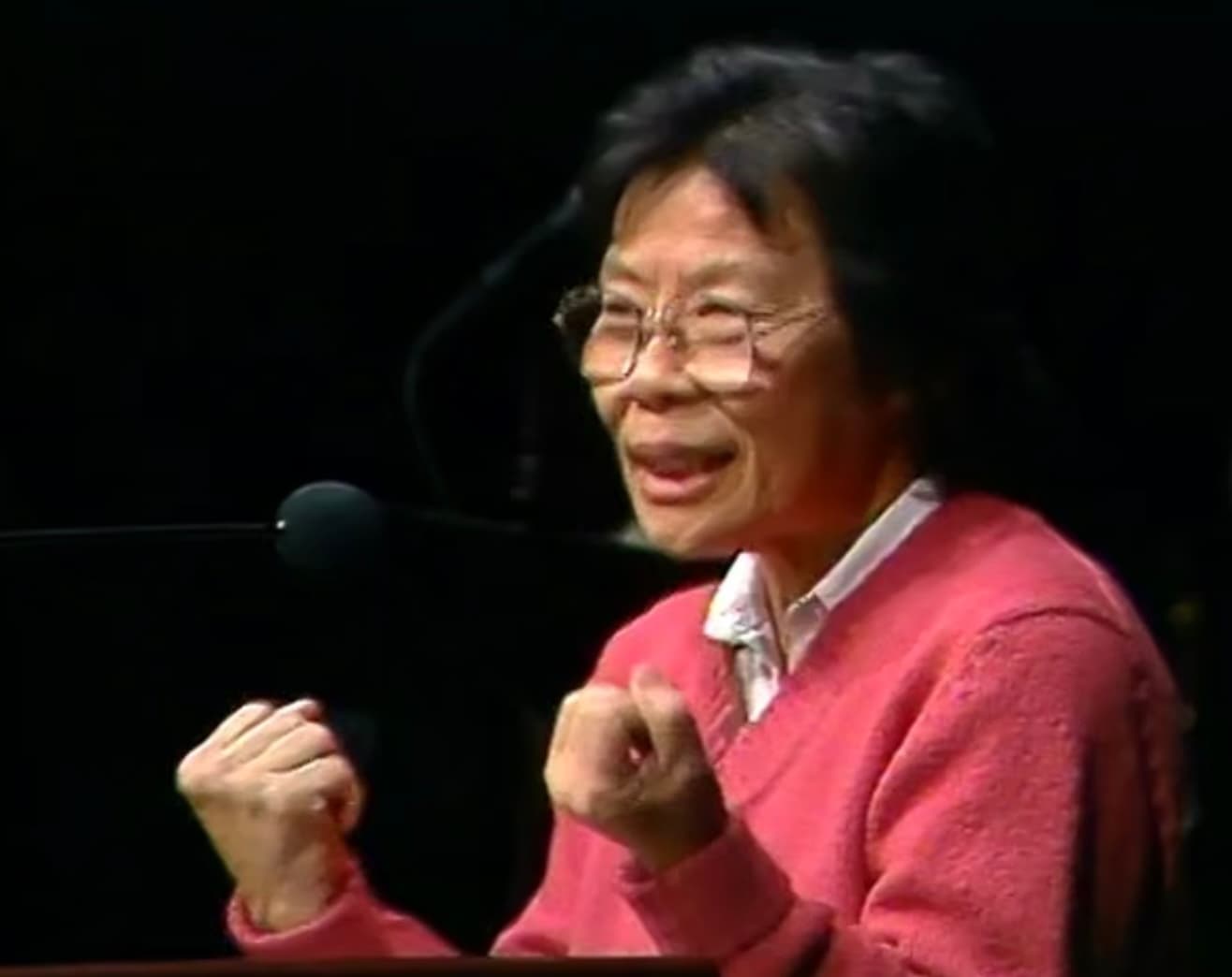A woman in a red cardigan speaks into a microphone while gesturing with both hands.
